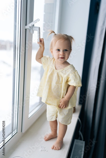 Fototapeta Vertical portrait of adorable toddler girl trying opening window. Concept of critical need for child safety measures to prevent home accidents and ensure secure environment for exploration.
