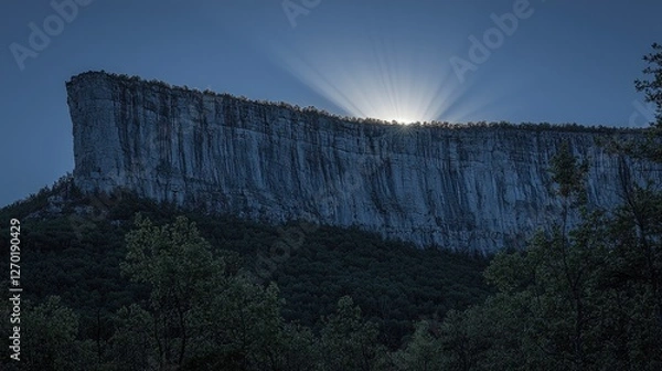 Fototapeta Majestic cliff at sunset with rays of light emerging behind, surrounded by lush forest