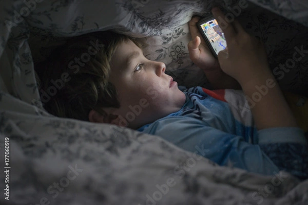 Obraz Young boy playing with cell phone under duvet