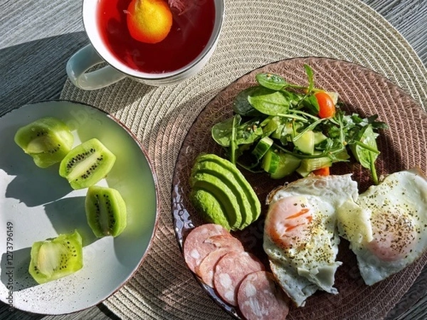 Fototapeta tasty healthy morning breakfast with a cup of coffee or tea. lots of food on the table with a green salad before starting the day. eggs with avocado and kiwi fruits. organic nutrition for health