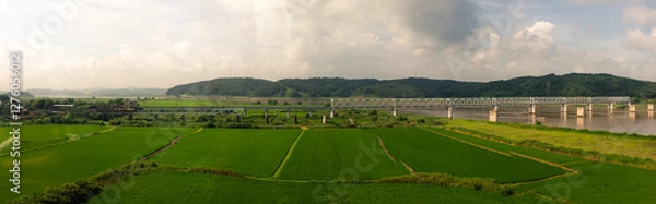 Fototapeta Scenic Green Fields With Flowing River And Steel Bridge Surrounded By Rolling Hills Under Partly Cloudy Sky