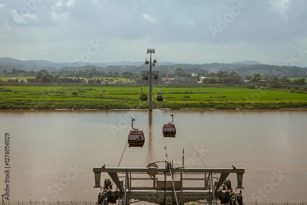 Fototapeta Scenic Cable Cars Gliding Over A Serene River With Green Fields And Rolling Hills In The Background