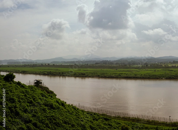 Fototapeta Watchtower Overlooking Serene River And Expansive Green Fields With Rolling Mountains Under Cloudy Sky