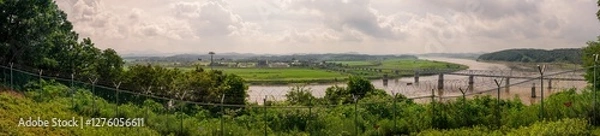 Fototapeta Expansive Panoramic View Of Green Fields And River With Bridges And Cable Cars Beyond A Barbed Wire Fence