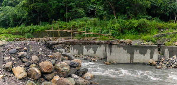 Fototapeta Unfinished bamboo bridge spanning rapid river with broken concrete supports amid lush green jungle scenery