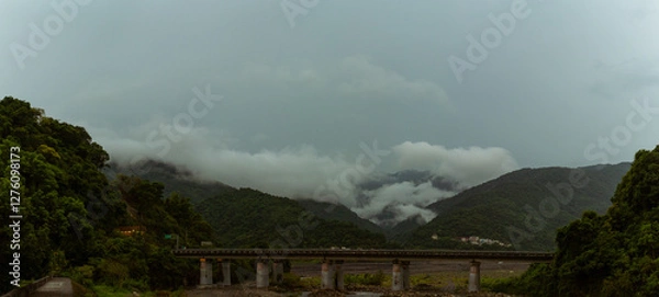 Fototapeta Dense mist rolling over lush green mountains with elevated bridge spanning river valley under cloudy sky
