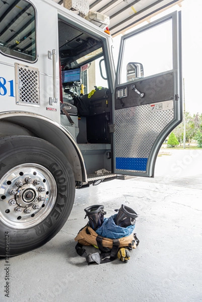 Fototapeta A fire truck with the door open and fire fighter boots and pants at the ready on the floor next to the truck door, inside a fire station with the bay door open ready to go out on a call. 