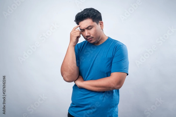 Fototapeta An Asian man in a blue t-shirt stands against a plain white background, pointing to his temple with a skeptical and questioning expression. His posture suggests deep thought, confusion, or curiosity