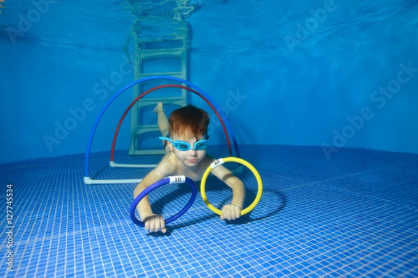 Obraz Baby training underwater in the pool and swims through hoops. The view from under the water. Landscape orientation