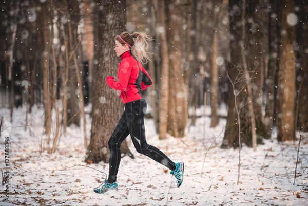Fototapeta Winter running exercise. Runner jogging in snow. Young woman fitness model running in a city park 