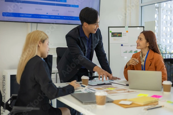 Obraz Three people are sitting at a table with a laptop open. One of them is laughing while the other two look on. The man in the middle is giving a presentation to the other two

