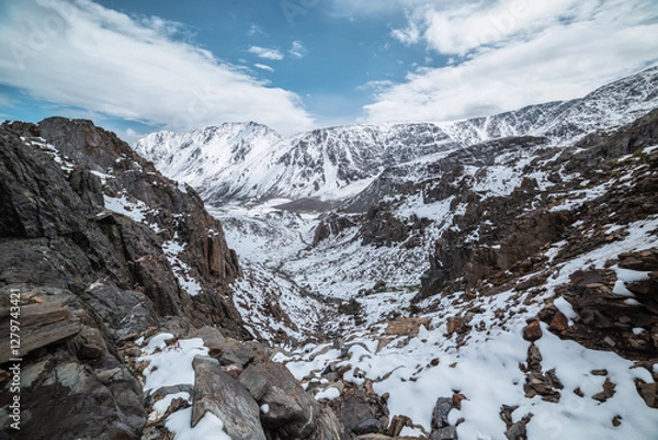 Fototapeta Descent along thin rocky gorge down into snow-covered alpine valley. Landscape from above to snowy stony couloir. White snow on stones in high mountains. Top view to snow-white steep slope among rocks