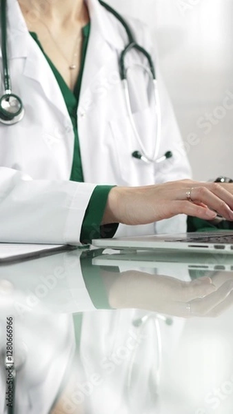 Fototapeta A female doctor in a green blouse under a medical coat is using technology in healthcare. Close-up of hands typing on a laptop on a glass desk in the clinic, vertical view. Medical concept
