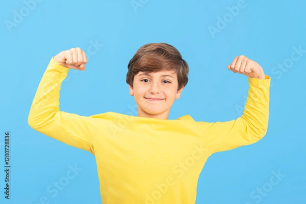 Fototapeta Boy raising arms with clenched fists, showing biceps, smiling face, yellow t-shirt, light blue background, winner gesture