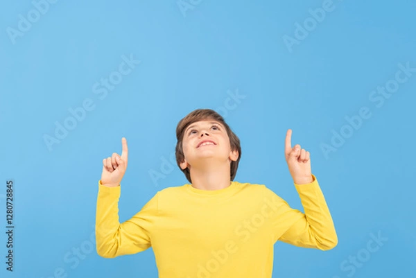 Fototapeta Charming young boy pointing and pointing with arms and fingers upwards in yellow sweater, showing something interesting and surprising, isolated on blue background