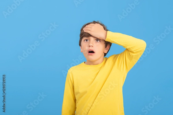 Fototapeta Young boy holding hands to head, amazed, surprised, surprised, confused, resigned, in yellow sweater, isolated on blue background. Concept of disaster
