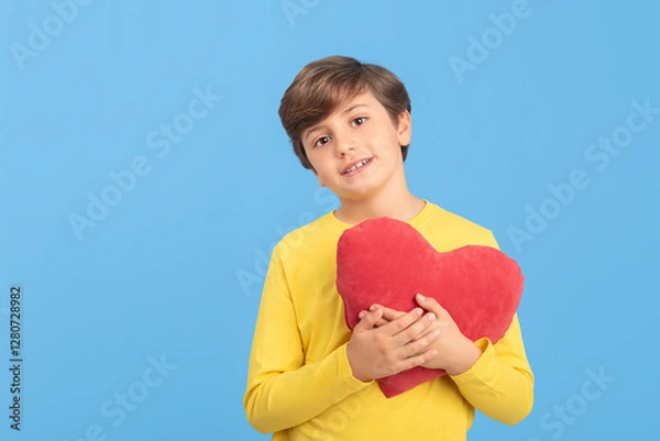 Fototapeta Charming young boy holding red heart in his arms in yellow sweater, isolated on blue background. Concept of love, compassion and good feelings