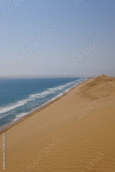Fototapeta Towering sand dunes cascade towards the ocean, shaping an otherworldly landscape in Sandwich Harbour. The meeting point of the desert and sea creates a mesmerizing, untouched natural beauty.