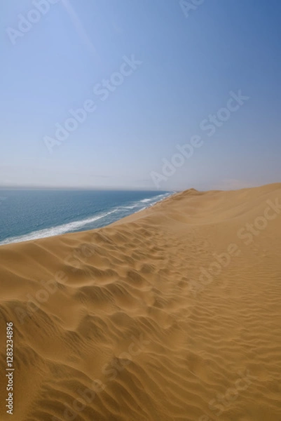 Fototapeta Majestic dunes of Sandwich Harbour meet the Atlantic Ocean, where golden sand stretches endlessly, merging with the deep blue sea under a vast sky. A breathtaking desert-coastline contrast.
