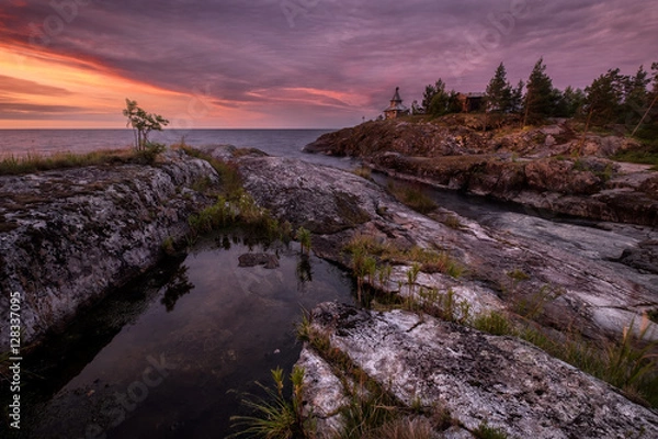 Obraz An island in the Ladoga lake with striped granite cliffs and small pond. Smooth surface of the pond reflects clouds lighted by a vivid sunrise. An orthodox church stands at cape of the island, Karelia