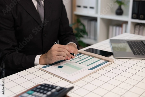 Fototapeta A man in a suit is holding a pen and looking at a piece of paper. He is focused and attentive, possibly reviewing some important information or making a decision
