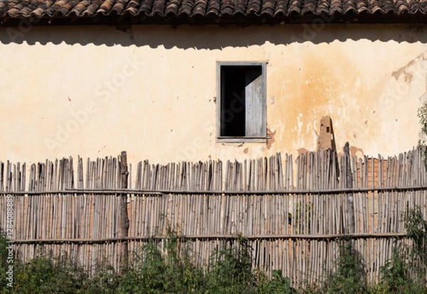 Fototapeta facade of old house with sunburned wall and old clay tiles with a single window half open and half in darkness with small fence made of bamboo pieces on a strong sunny day that produces strong shadows