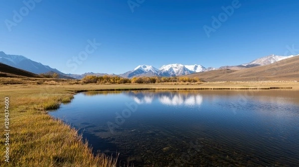 Fototapeta A serene landscape featuring a calm lake reflecting snow-capped mountains under a clear blue sky, surrounded by lush grassy terrain.