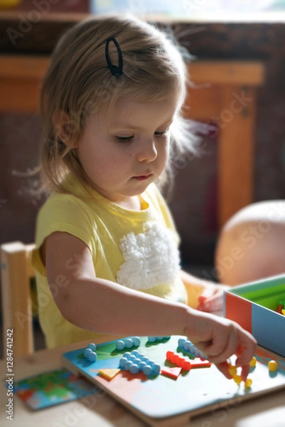 Fototapeta A little girl of three years old plays with an educational game, a multi-colored mosaic, laying it out on a substrate. Development of preschool children. Educational toys for kids. Fine motor skills.