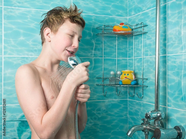 Fototapeta Young boy with red hair and naked torso holding a watering can shower and sings on the background of blue tiles and bottles of shampoo