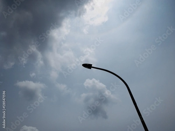 Fototapeta Silhouette of a curved streetlight against a cloudy sky with sunlight filtering through. A moody and dramatic scene showcasing urban minimalism and contrast between light and dark.