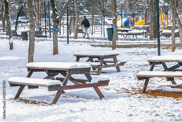 Fototapeta A serene winter scene in a park with snow-covered picnic tables and a playground in the background.