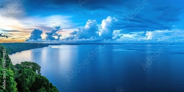 Fototapeta Serene sunset over a vast, blue river, lush green rainforest visible on the left, fluffy white clouds in a vibrant blue sky. Peaceful, tranquil