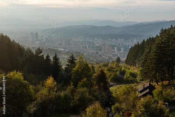 Obraz Mountain top view of city at sunset. A sprawling cityscape partially covered in light fog, illuminated by the warm hues of an early autumn sunset, creating a serene and atmospheric scene.