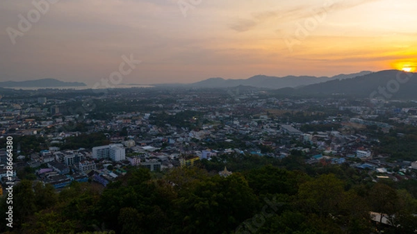 Fototapeta A scenic view of a town nestled among lush green mountains at sunset, with a temple structure perched on the hillside and a vibrant orange sky creating a warm, tranquil atmosphere.