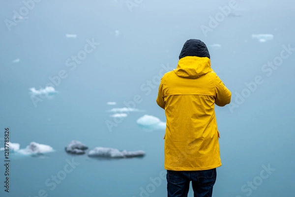 Obraz A man with a wet yellow raincoat on a rainy day standing in front of a blue laggoon with ice floes
