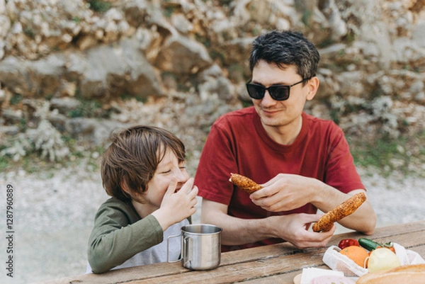 Fototapeta Close-up view of father hugs his school boy son on a family picnic. Child kid and his dad taking a rest and enjoying a picnic while hiking. Boy smiles and bites the bread donut on a picnic.