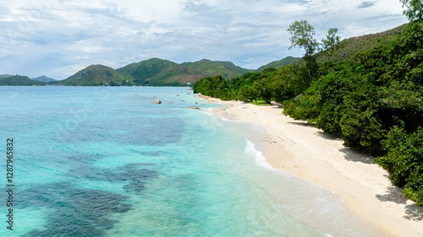 Fototapeta A beautiful white sandy beach with calm turquoise waters, framed by lush green trees and distant rolling hills. Praslin, Seychelles.