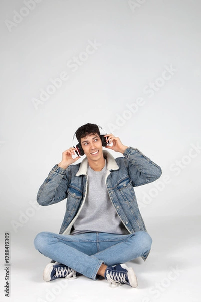 Obraz Chico joven vestido de vaqueros y camiseta blanca sujetando auriculares y mirando a un lado sonriendo. Fondo blanco. Concepto de música, tecnología y publicidad.