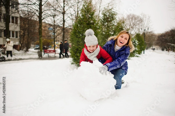 Fototapeta Mother with daughter of younger school age build a snowman in the yard.