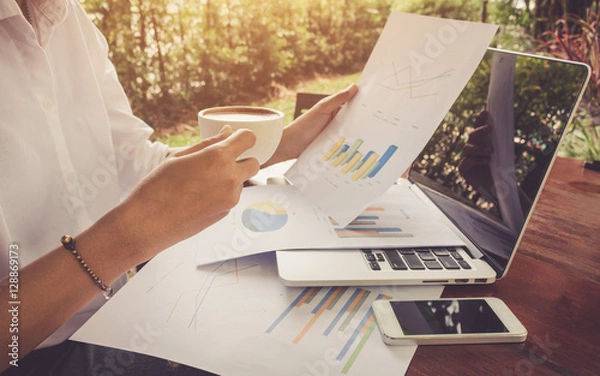 Obraz Biznes kobieta analizuje dokument wykresu z laptopa i trzyma ton kawy rocznika.