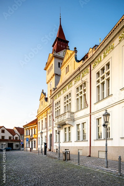 Fototapeta The historic Town Hall in Stara Boleslav stands proudly in the early morning light, showcasing its remarkable architecture along a quiet cobblestone street lined with trees.