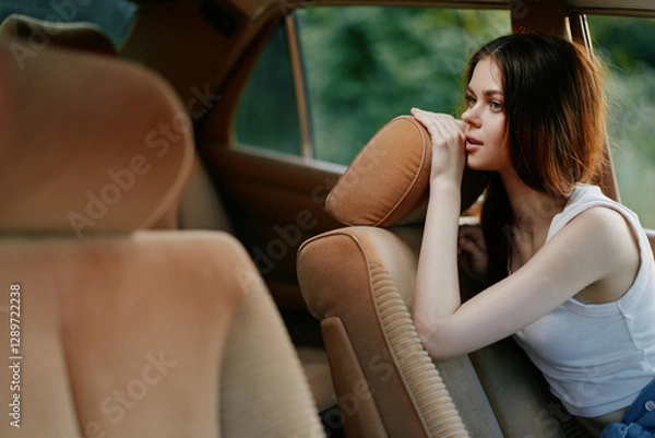 Fototapeta Thoughtful young woman in a car contemplating life, wearing a casual white tank top, with a serene expression and blurred green background emphasizing tranquility.