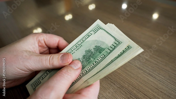 Fototapeta A pair of hands is seen counting several one hundred dollar bills at a wooden table. The setting is bright with natural light coming through nearby windows.