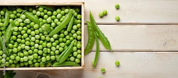 Obraz Fresh green peas in a wooden box on a rustic white table with scattered peas and pea pods highlighted against a clean background.