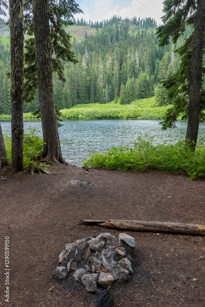 Fototapeta This serene lakeside campsite showcases a stone fire pit nestled under towering pine trees, with vibrant greenery and a calm body of water in the background, perfect for relaxation.