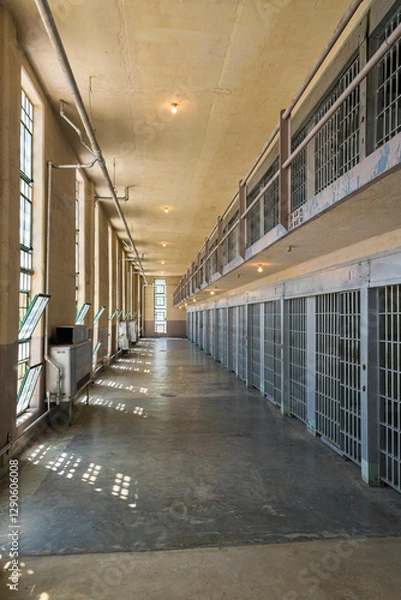 Fototapeta A long, empty corridor inside a modern prison building, featuring multiple cells with barred windows and bright lights, highlighting the building's architectural design and purpose.