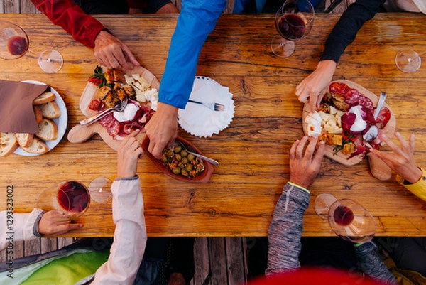 Fototapeta Friends sharing a rustic meal together outdoors - group of people enjoying a shared meal with wine, cheese, bread, and charcuterie on a wooden table