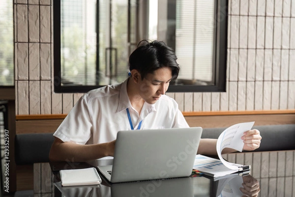 Fototapeta Young professional analyzing documents while working on laptop in modern office, focusing on data and project details.