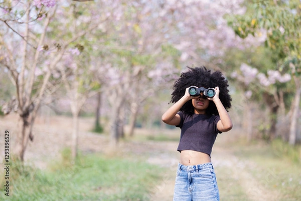 Obraz A young woman with voluminous curly hair explores nature using binoculars. Dressed in a black crop top, she stands on a dirt path surrounded by blooming trees.