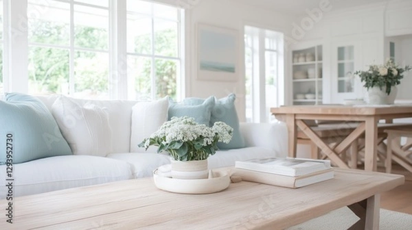 Obraz A living room with a white couch, a coffee table, and a dining table. The coffee table has a vase with flowers on it, and there are books on the table. The room has a clean and organized look